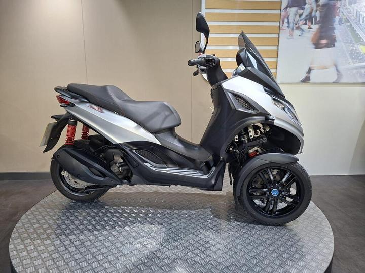 Piaggio MP3 300 Sport (2022) A silver three-wheeled scooter displayed on a circular platform with a textured surface, set against a neutral-colored backdrop. The scooter features a sleek design, a comfortable seat, and red suspension springs.