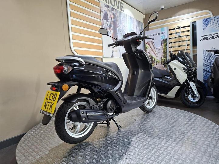 A black scooter parked on a display stand with a clear view of its rear and side, featuring a license plate that reads "LG18 NTK". In the background, other scooters are visible along with promotional materials related to urban mobility.