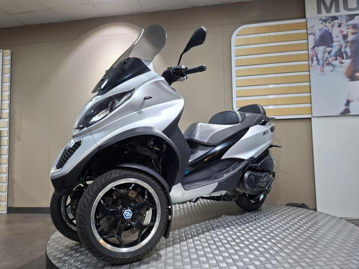 A silver three-wheeled scooter displayed on a textured platform, featuring a sleek design with a windshield and sporty wheels. The background includes beige walls and a large image of people walking.