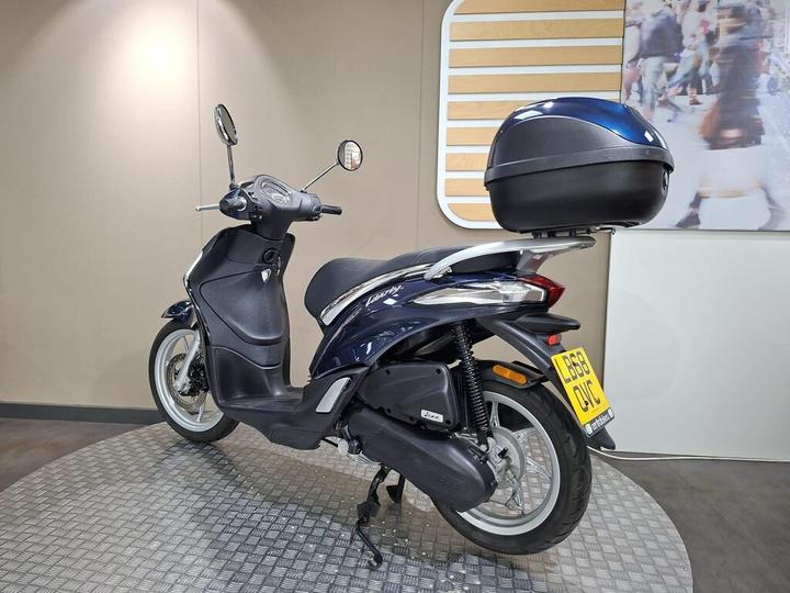 A dark blue scooter with a black top case is displayed on a circular platform against a neutral background.