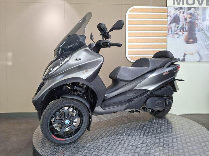 A grey three-wheeled scooter displayed on a circular platform with a textured surface, featuring a large windshield, sporty design, and a prominent logo on the front wheel. In the background, there is a blurred image of shoppers.