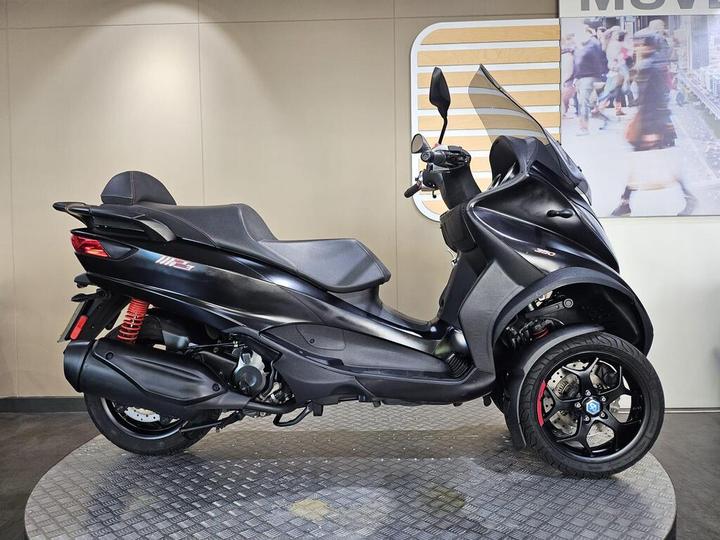 A black three-wheeled scooter displayed on a circular rubber mat, featuring a sleek design, sporty details, and a windshield, set against a neutral background with an image of people in motion.