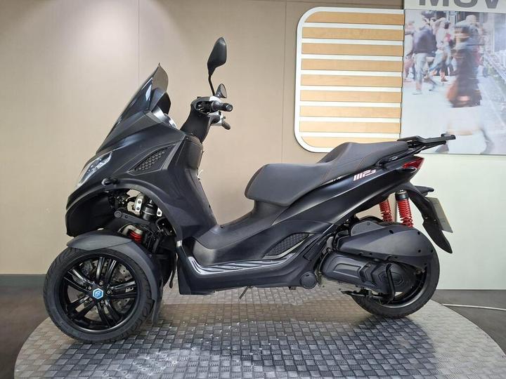 A black three-wheeled scooter displayed on a circular platform against a neutral background. The scooter features a streamlined design, with a prominent windshield, ergonomic seat, and distinctive red suspension springs.