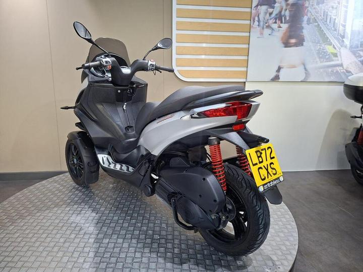A modern scooter showcased in an indoor setting, featuring a sleek silver and black design, dual rear suspension with red springs, and a yellow license plate. The scooter has a unique body shape and is positioned on a textured floor.
