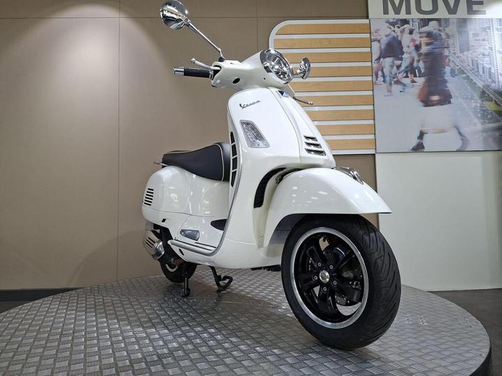 A white vintage-style scooter is positioned on a textured circular platform, showcasing its sleek design and black wheels. The background features a light-colored wall and a large image of people walking in a city scene.