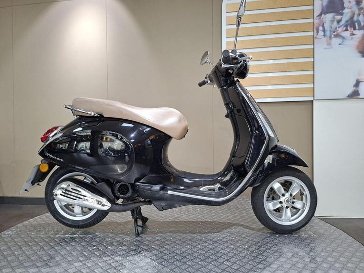 Vespa Primavera 50 (2017) A black scooter with a brown seat is positioned on a textured platform, set against a neutral background with a hint of people in motion visible in a nearby display.