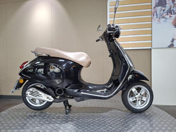 Vespa Primavera 50 (2017) A black scooter with a brown seat is positioned on a textured platform, set against a neutral background with a hint of people in motion visible in a nearby display.