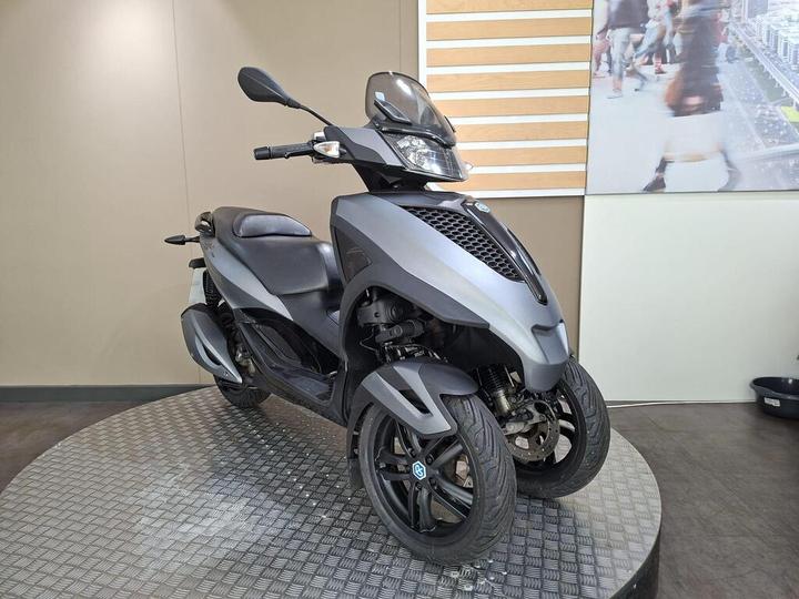 A grey three-wheeled scooter displayed on a circular platform in an indoor environment, featuring a sleek design and modern styling.