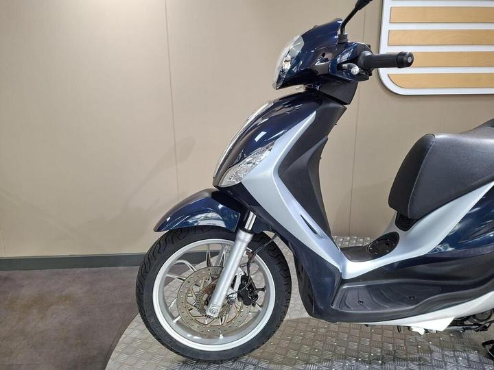 A side view of a blue and silver scooter, showcasing its front wheel, suspension, and part of the seat against a neutral background.