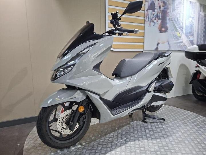 A modern gray scooter with a sleek design is displayed in a showroom, featuring prominent headlights, a sporty front fairing, and a comfortable seat. The background includes a light-colored wall and a blurred image of pedestrians.
