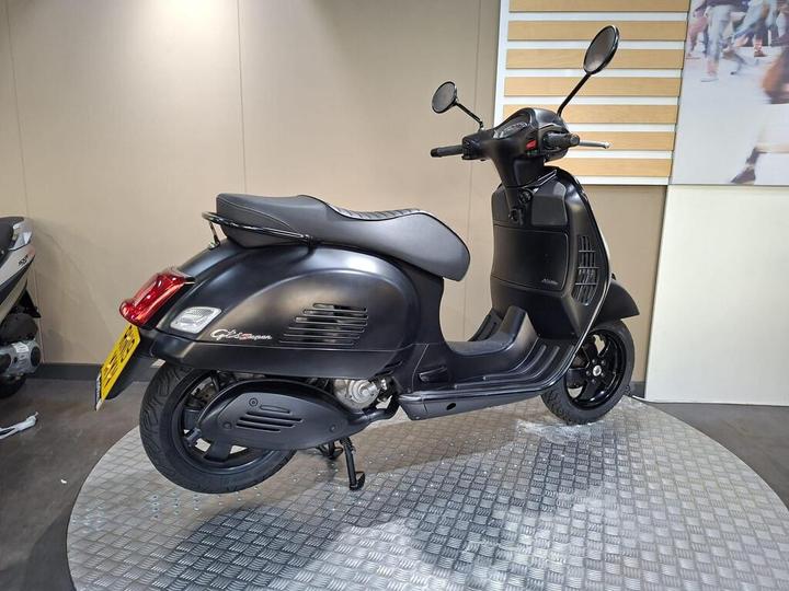 A sleek black scooter displayed on a circular traction sheet in a showroom, with its rear side visible, featuring a modern design and chrome accents.