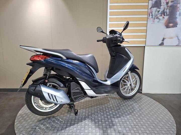 A sleek blue and silver scooter stands on a checkered platform, angled to show its side profile. The background features a neutral wall and a large image of people walking.