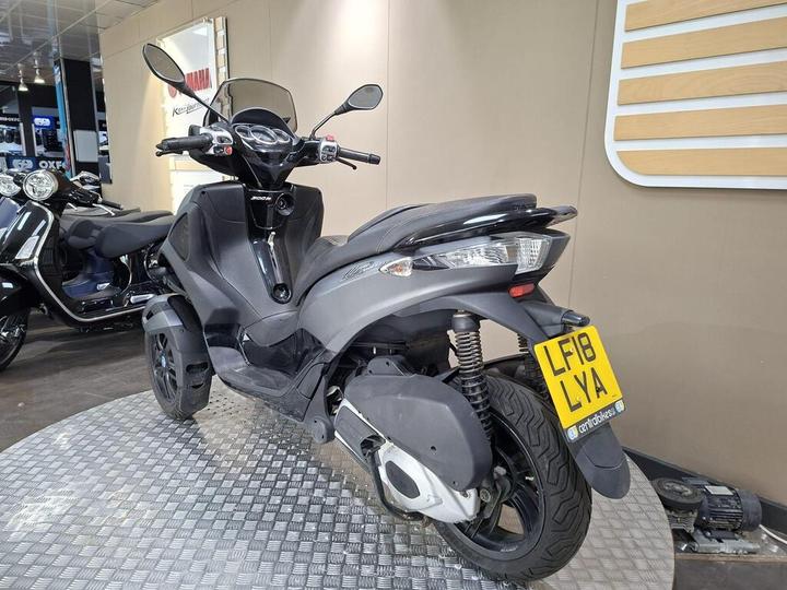 A black scooter with a unique design parked on a checkered floor, displaying the rear and side view. The vehicle's registration plate reads "LF18 LVA." In the background, another scooter is partially visible against a beige wall.