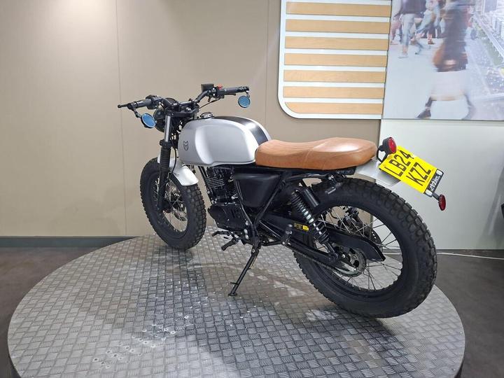A custom motorcycle with a silver gas tank, black chassis, and brown leather seat is displayed on a metallic rotating platform in a modern showroom.