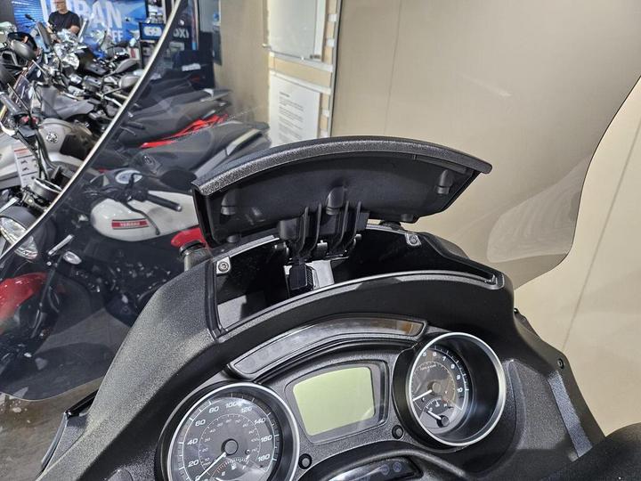 Close-up of a motorcycle dashboard with an open compartment, displaying gauges and dials. In the background, several motorcycles are visible, along with a reflective surface.