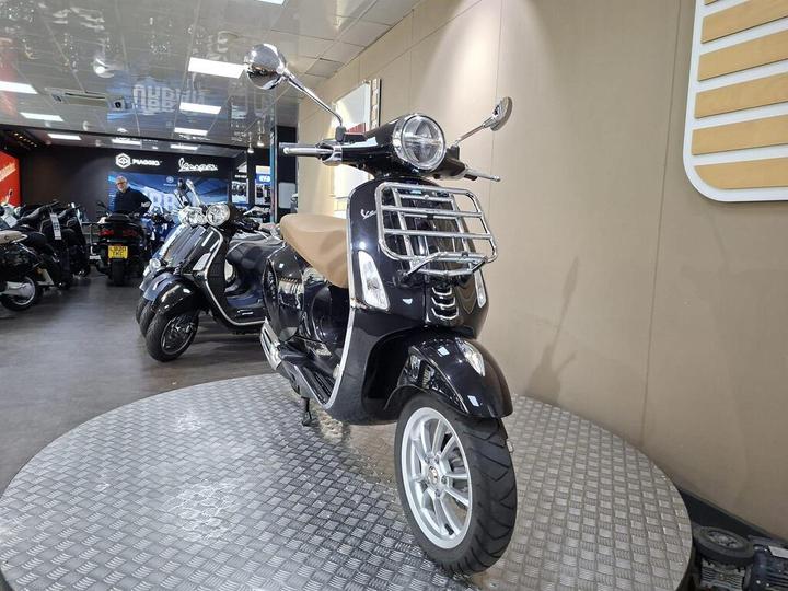 A glossy black scooter displayed on a circular platform in a showroom, featuring a brown seat, chrome accents, and a modern design. Other scooters are visible in the background.