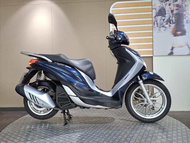 Piaggio Medley 125 (2018) A sleek blue scooter is displayed against a neutral wall backdrop, highlighting its design features, including a comfortable seat and alloy wheels.