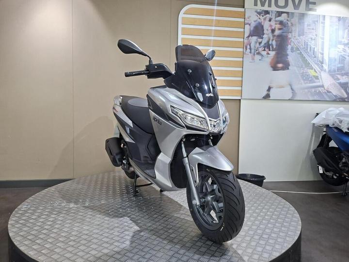 A sleek silver scooter is displayed on a circular platform, with a modern interior background featuring branding and a blurred image of pedestrians.