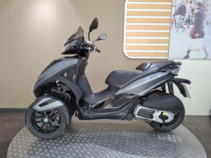A black and silver three-wheeled scooter displayed on a round metal platform, with a modern backdrop featuring a blurred image of people walking.