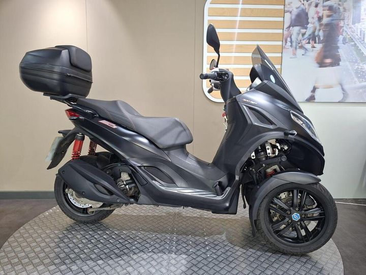 Piaggio MP3 300 Sport 2020 A black three-wheeled scooter is displayed on a textured circular platform, featuring a sleek design, a rear storage box, and red suspension springs. The background includes a softly lit wall with a modern pattern and a blurred image of people.