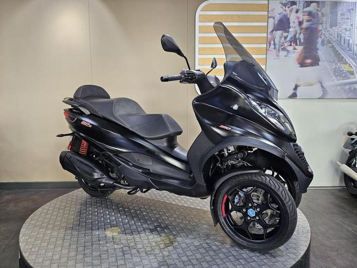 A black three-wheeled scooter displayed on a rotating pedestal, featuring a sleek design, large windshield, and sporty wheels. The background shows a blurred image of people walking.