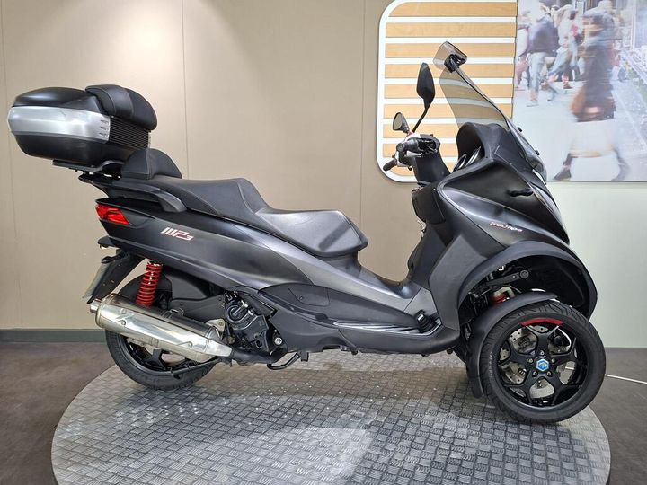 Piaggio MP3 500 HPE LT Sport 2020 A sleek black three-wheeled scooter with a large rear storage compartment, displayed on a circular platform with a textured surface. The scooter features sporty red rear suspension and distinctive wheel designs.