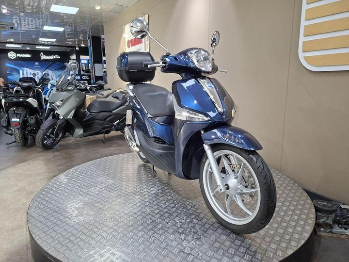A blue scooter displayed on a circular platform in a motorcycle showroom, with other scooters and motorcycles visible in the background.