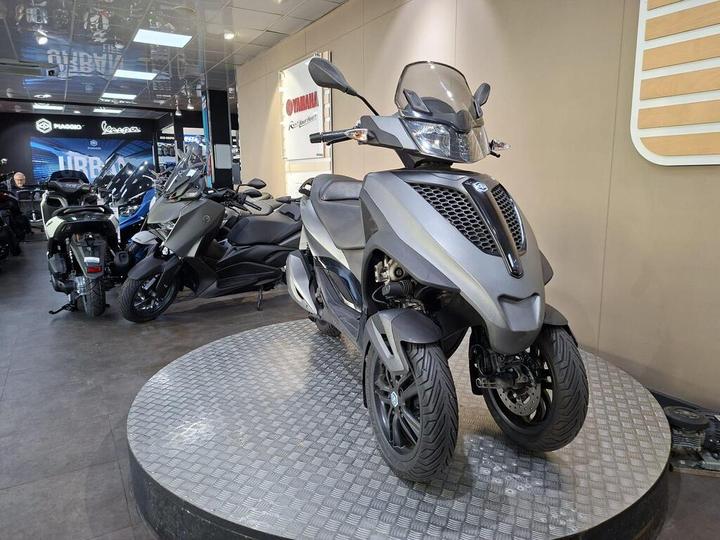 A three-wheeled scooter displayed on a circular platform in a motorcycle dealership, with a sleek gray design and front protective windscreen. Other motorcycles and scooters in the background are partially visible.