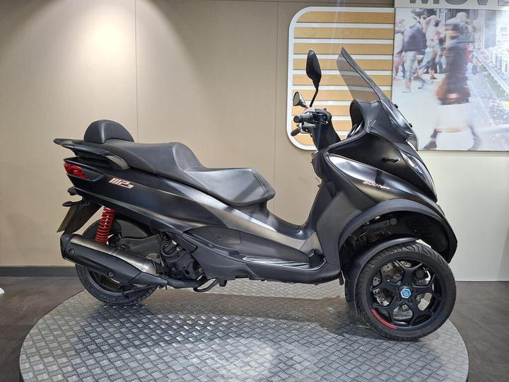 Piaggio MP3 500 Sport (2019) A black three-wheeled scooter with a streamlined design is displayed on a circular metallic platform against a neutral background.