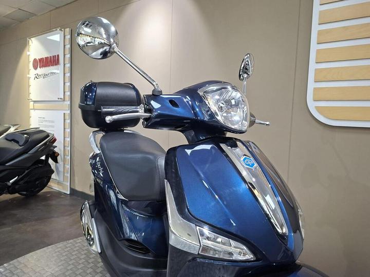 A close-up view of a blue scooter with a shiny front headlight and rearview mirrors, displayed in a showroom with a Yamaha logo visible in the background.