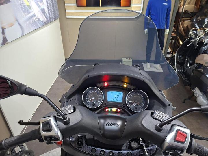 View from the handlebars of a scooter, featuring a digital speedometer displaying 60 km/h, various indicator lights, and a translucent windscreen, with a blurred background showing motorcycle gear and apparel.