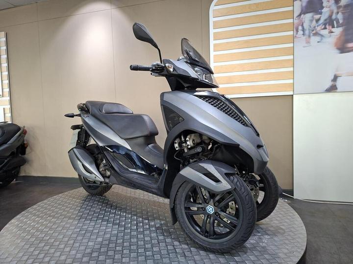 A sleek, gray three-wheeled scooter displayed on a circular platform in a modern showroom, featuring a streamlined design and a black seat. Soft lighting highlights its contours against a simple background.