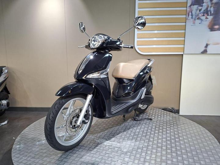 A sleek black scooter with a brown seat and chrome accents, positioned on a circular textured surface, set against a neutral wall with a partially visible image of people walking in the background.