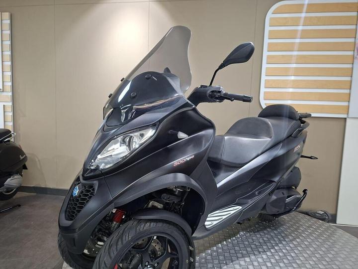 A sleek black three-wheeled scooter with a transparent windshield, parked indoors against a beige wall, showcasing its modern design and dual seating.