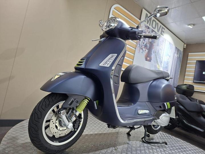 A blue modern scooter displayed in a showroom, featuring a sleek design, round headlight, and chrome details. The scooter is positioned on a metallic platform, with a blurred background showcasing other vehicles and a wall sign that reads "MOVE."