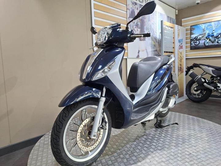 A blue and white scooter is displayed in a showroom setting, featuring a sleek design, front fender, and chrome accents. In the background, wall graphics and another motorcycle can be seen.