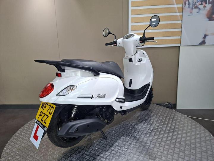 A white scooter is displayed in a showroom, showcasing its rear and side view. The scooter has a prominent red taillight and an "L" plate, indicating learner status, with a registration plate visible. The background features a neutral-colored wall and a display panel.