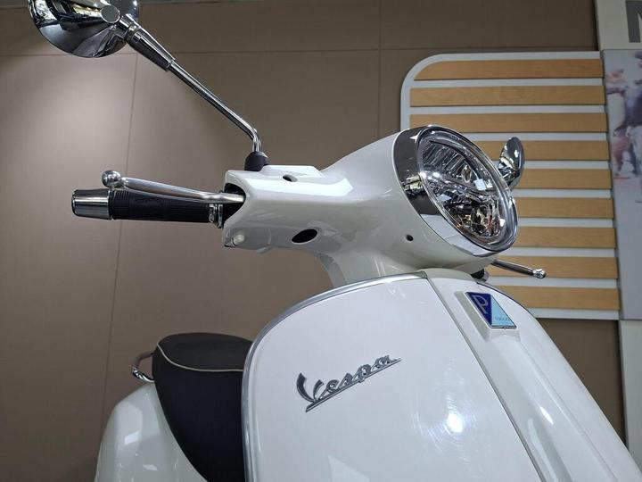 A close-up view of a white Vespa scooter's handlebars, showcasing the chrome mirror, headlight, and detailed logo on the front, with a blurred background.
