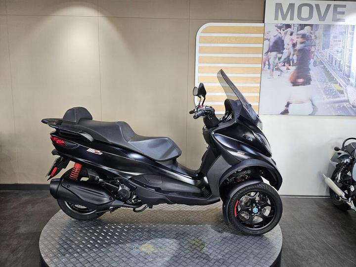A sleek black three-wheeled scooter is displayed on a circular platform, showcasing its modern design, with a transparent windscreen and red accents. The background features a wall graphic with the word "MOVE."