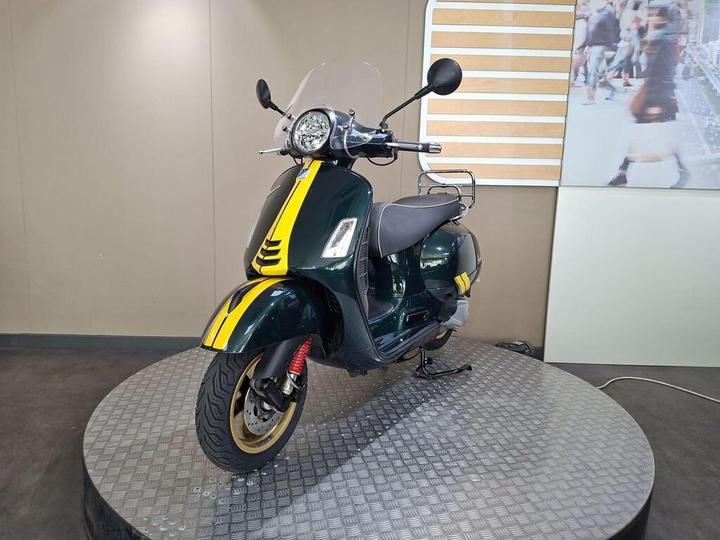A green scooter with yellow stripes and a windscreen, displayed on a circular metallic platform in a modern exhibit setting.