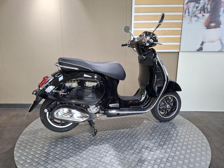 A black scooter displayed on a circular, textured platform in a showroom setting, featuring chrome accents and a glossy finish.