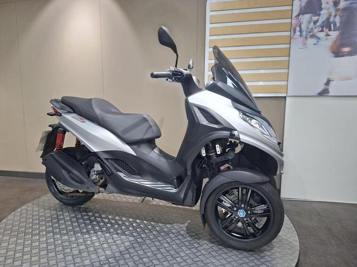 A sleek silver three-wheeled scooter displayed on a circular platform against a neutral background, showcasing its modern design and features.