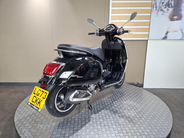 A black scooter displayed on a circular platform, viewed from the rear, showcasing its sleek design and rear license plate. The background features a neutral wall and an abstract image.