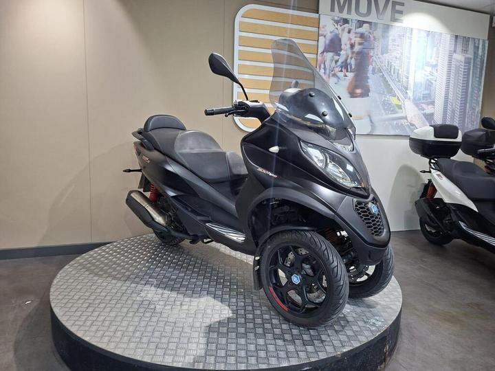 A black three-wheeled scooter with a windshield and black seats displayed on a circular platform, with a modern backdrop featuring city imagery.