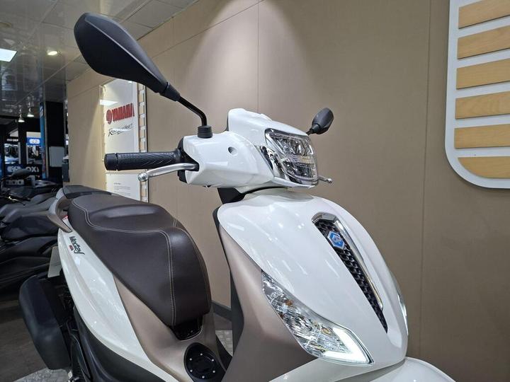 A close-up of a white scooter showcasing the handlebars, mirror, headlight, and a part of the seat, set against a neutral background in a showroom.