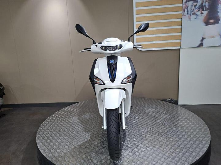 A white electric scooter displayed on a circular platform, viewed straight on. The scooter features a sleek design with a prominent front headlight, mirrors, and a textured tire. Neutral-colored walls and a partially visible image of people in the background.