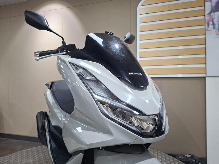 A close-up of a white Honda scooter with a sleek design, showcasing its front, headlight, and handlebars against a neutral background.