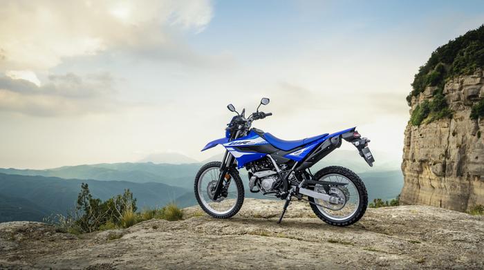 A blue dirt bike parked on a rocky outcrop with mountains and a cloudy sky in the background.