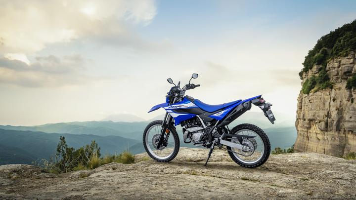 A blue dirt bike parked on a rocky outcrop with mountains and a cloudy sky in the background.