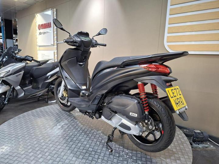 A sleek black scooter is displayed on a circular metal platform, showcasing its rear view. It features red shock absorbers and is positioned next to a silver motorcycle. The background includes a Yamaha promotional sign.